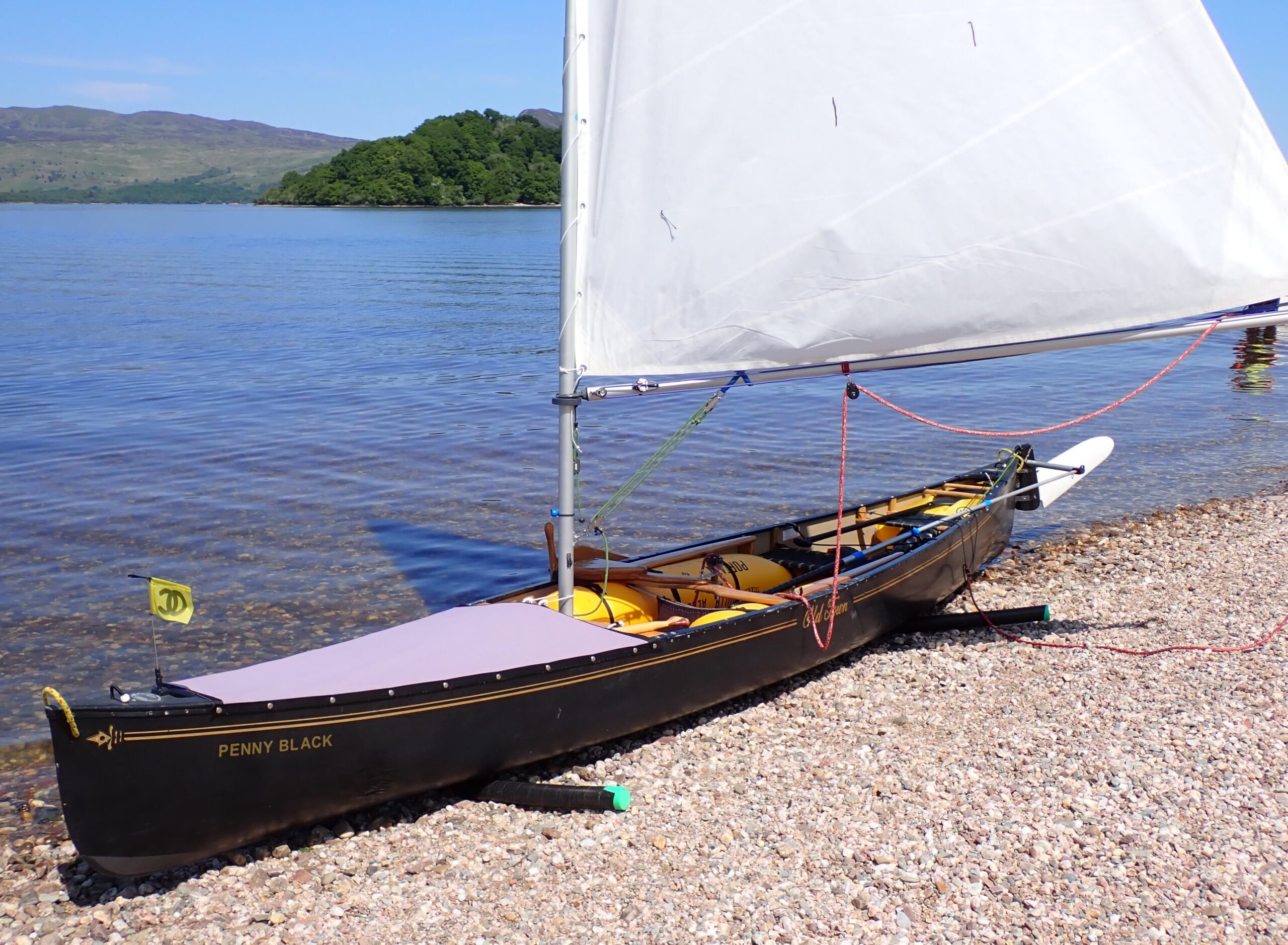 black sailing canoe on stony beach by loch with calm water and blue sky